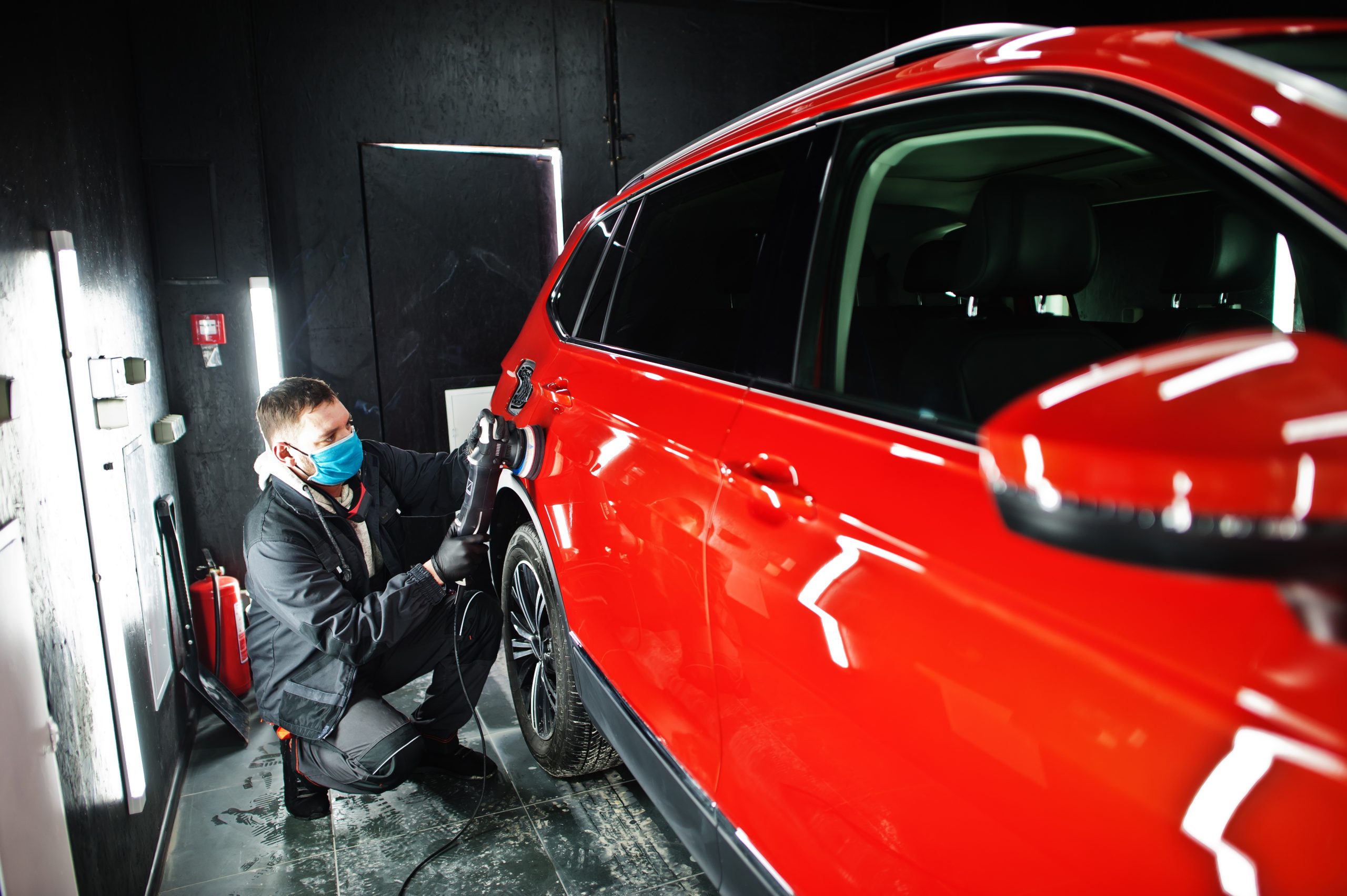 Car detailing concept. Man in face mask with orbital polisher in repair shop polishing orange suv car.